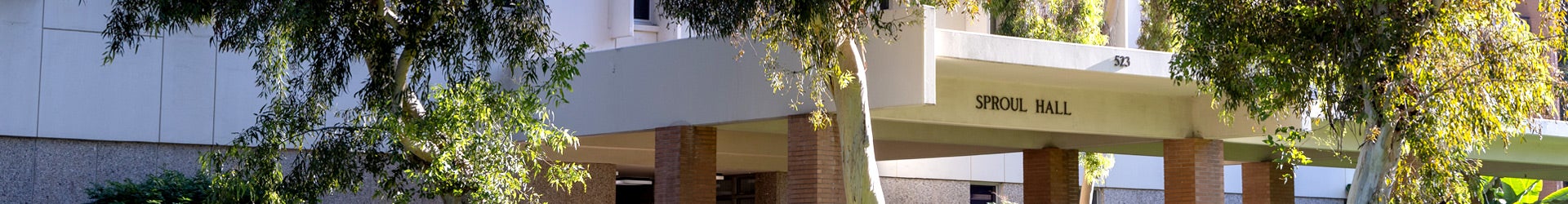 An exterior view of Sproul Hall on the UC Riverside campus. The building features cream-colored architecture and brick pillars partially framed by green leafy trees.