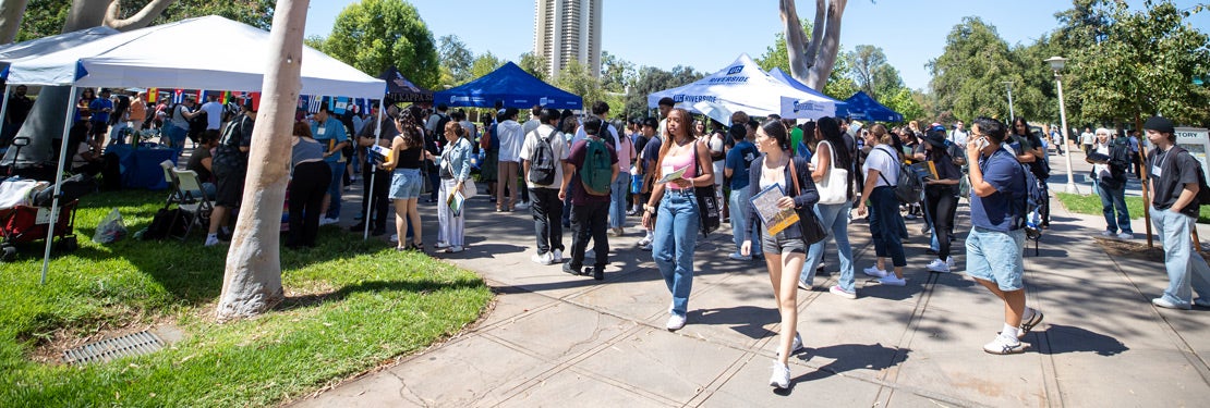A diverse group of students explores a vibrant UC Riverside transfer student event on campus. Under sunny skies, attendees walk along a paved walkway lined with informational tents to connect with university representatives.