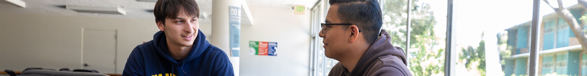 Two male UCR students wearing hoodies sit indoors near a large window and have a friendly conversation.