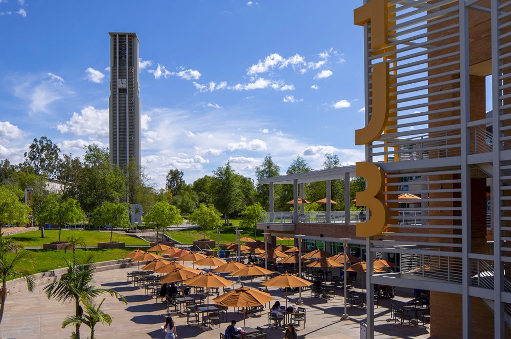 The UCR Bell Tower on a sunny day, overlooking an outdoor plaza with students studying under many orange umbrellas.